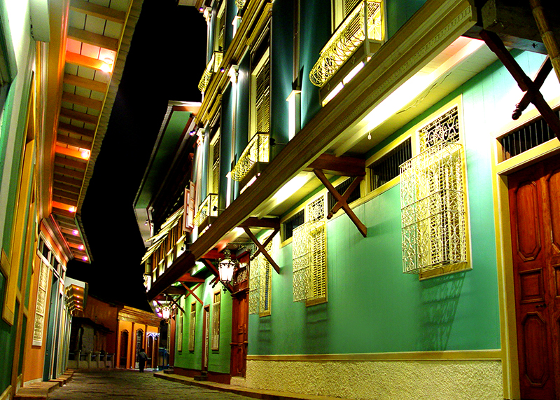Colorful historic buildings with decorative window grilles line an illuminated cobblestone street at night