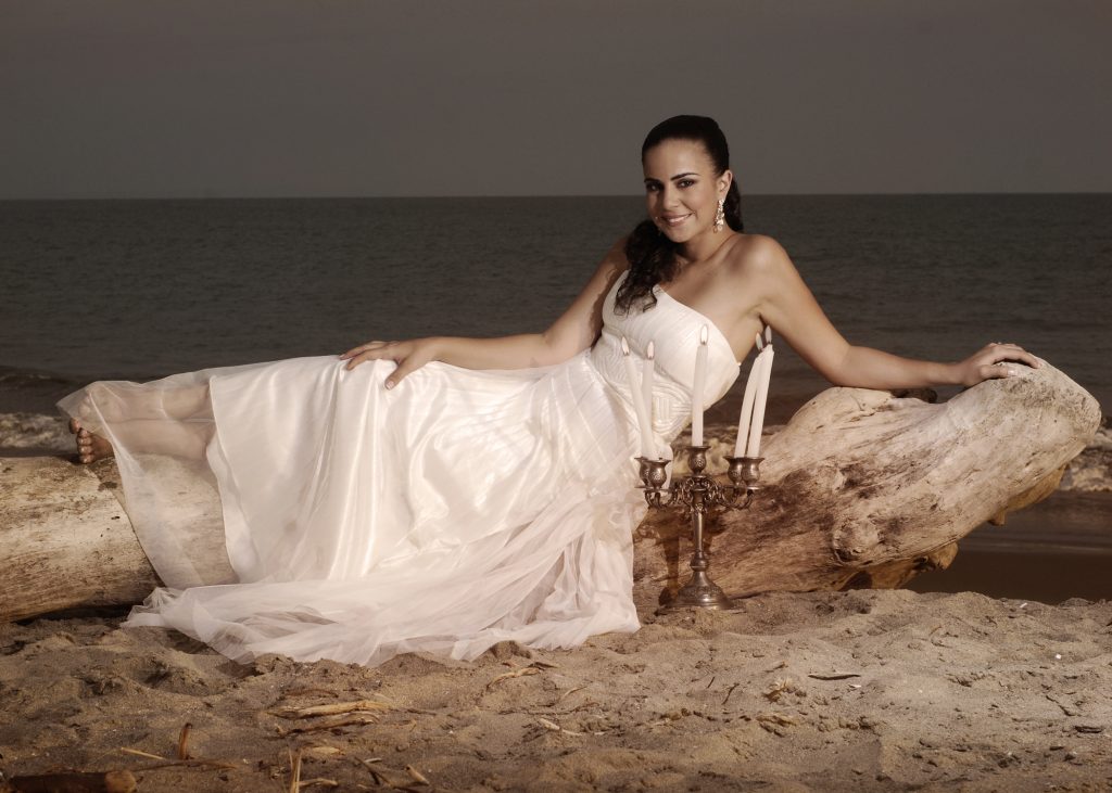 A smiling bride in a white wedding dress reclines on driftwood on a beach beside a candelabra with lit candles