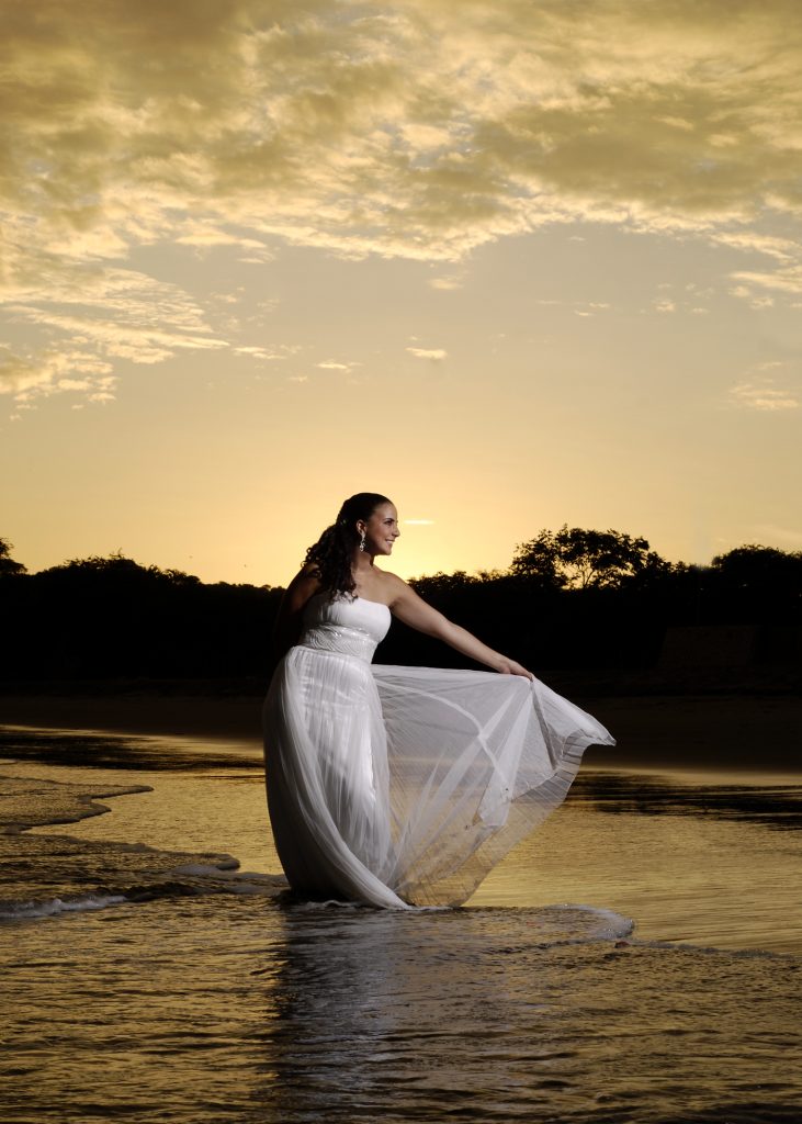 A bride in a white wedding dress holds up her flowing skirt while standing in shallow ocean water at sunset