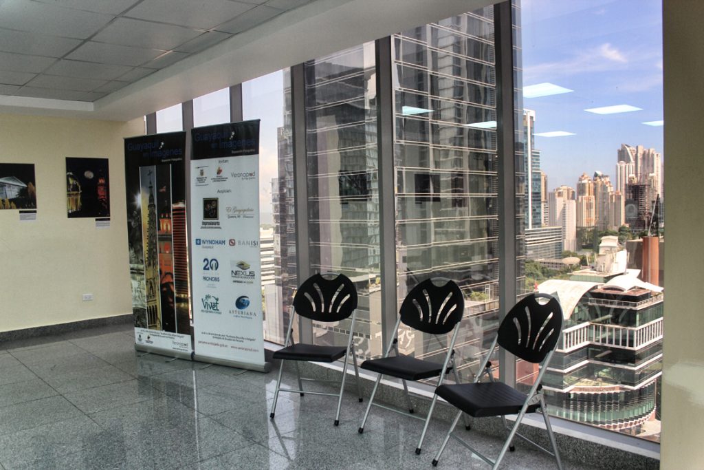 Three black chairs and Guayaquil en Imágenes banners stand by floor-to-ceiling windows showing a city skyline