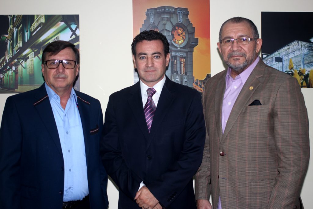 Three men in suits stand side by side in front of framed photographs displayed on a wall
