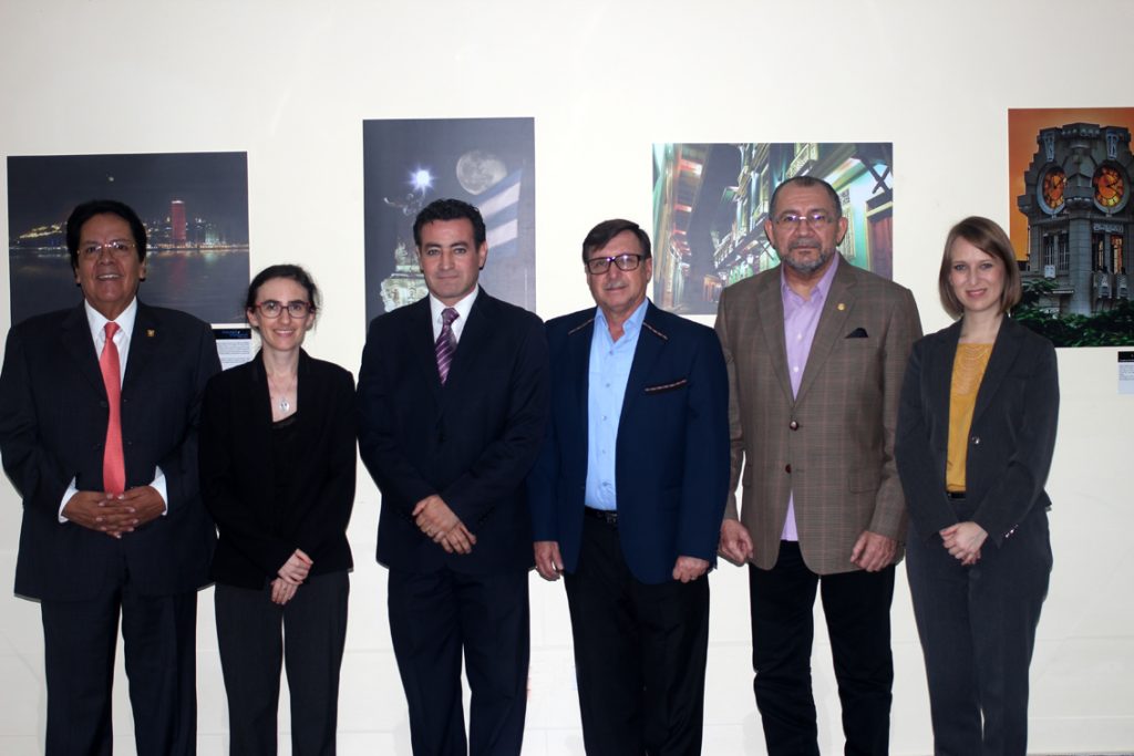 Six people in formal attire stand in a row in front of photographic prints of Guayaquil on a white wall