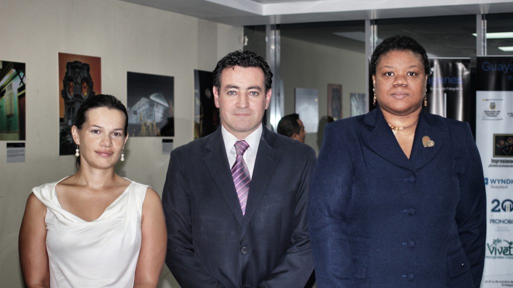 Three people stand together in formal attire in front of framed photographs at an exhibition