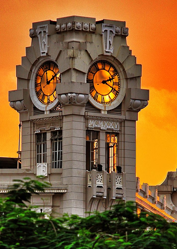 An ornate clock tower with illuminated faces and the Roman numerals MCMXXV stands against an orange sky