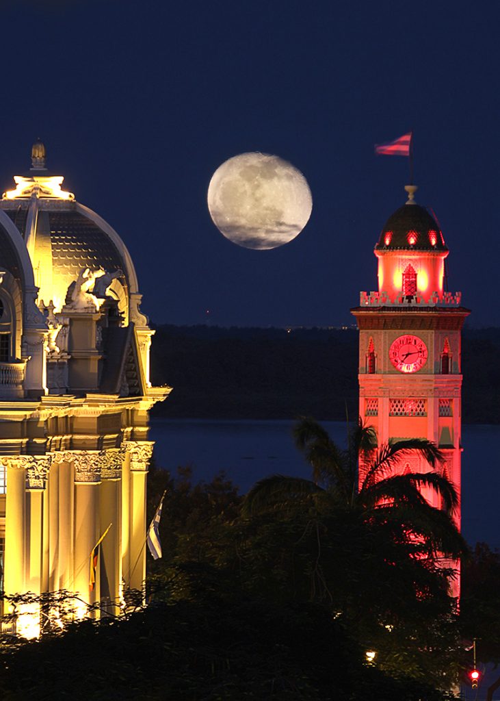 A full moon shines in the dark night sky between an illuminated golden building dome and a red clock tower
